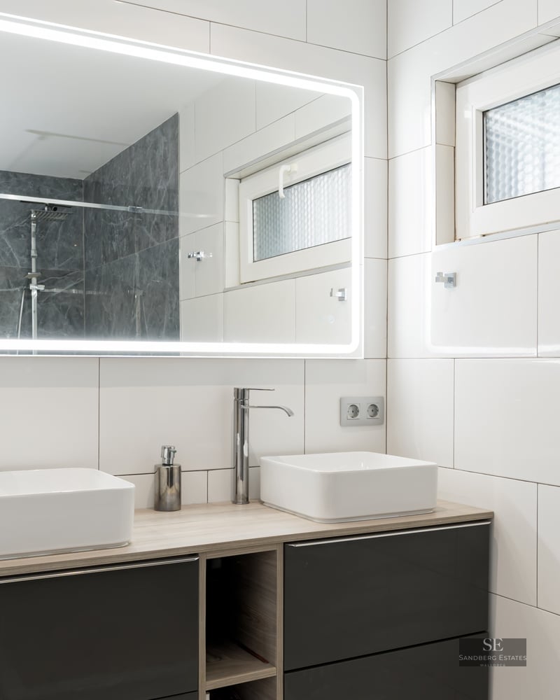 Salle de bain moderne avec deux vasques carrées, un grand miroir éclairé et des murs en carrelage blanc.