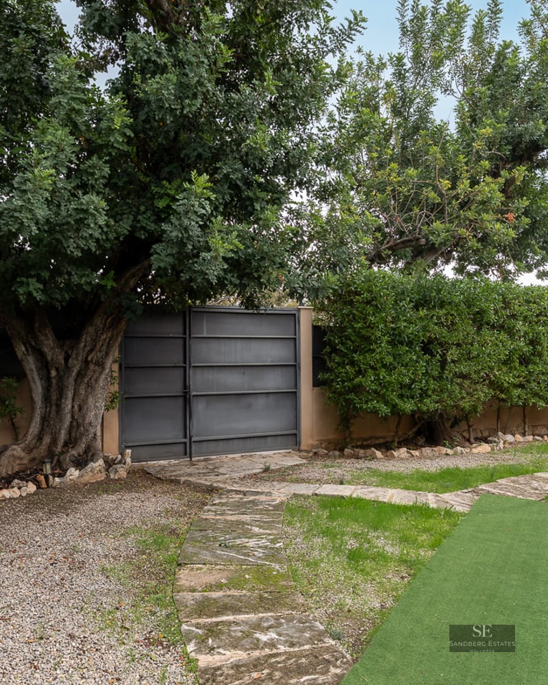 Private garden featuring a stone path, high hedges, a large mature tree, and a metal gate under natural daylight.