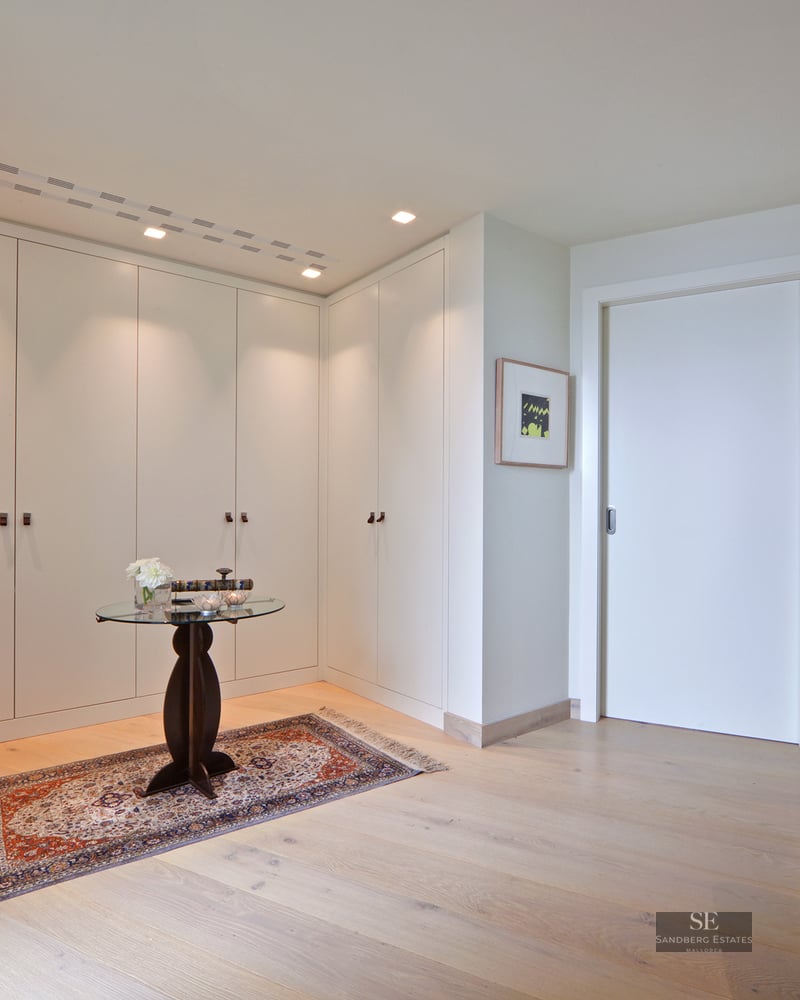 Bright entrance hall with white built-in closets, light wood floors, a decorative rug, and a glass side table.