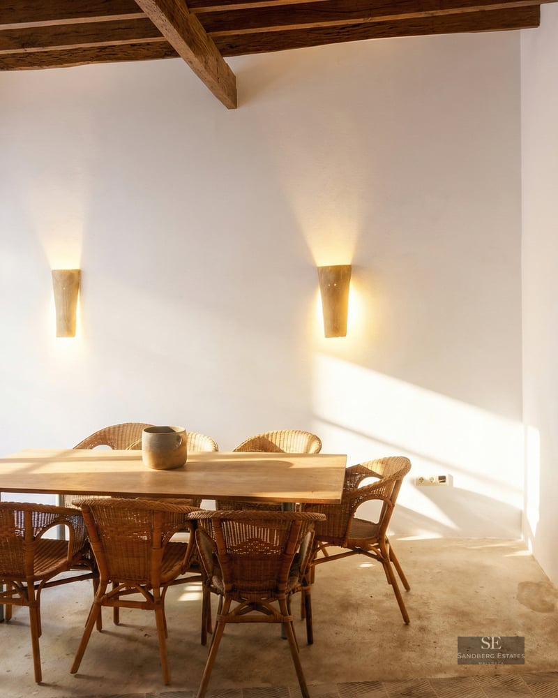 A wooden dining table with wicker chairs in a white room featuring dark ceiling beams and warm sunlight.