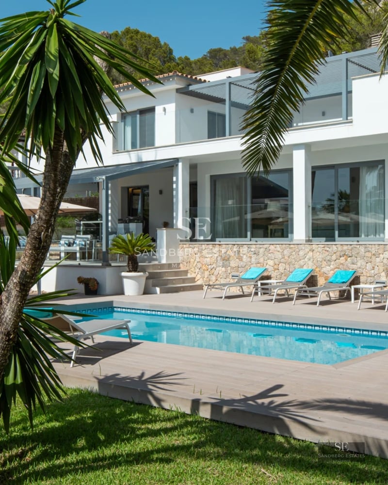 Large swimming pool surrounded by lush greenery, with a terrace and views of a modern and minimalist design house.