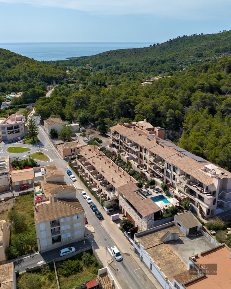 Drone shot of Mediterranean apartment buildings with a swimming pool, surrounded by green forest and sea horizon.