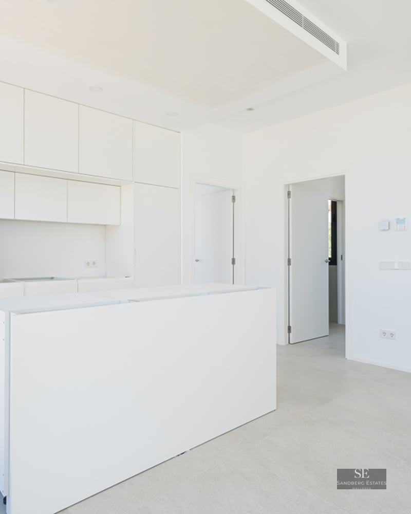 Bright white modern kitchen with central island, integrated appliances, and a wooden staircase leading upstairs.