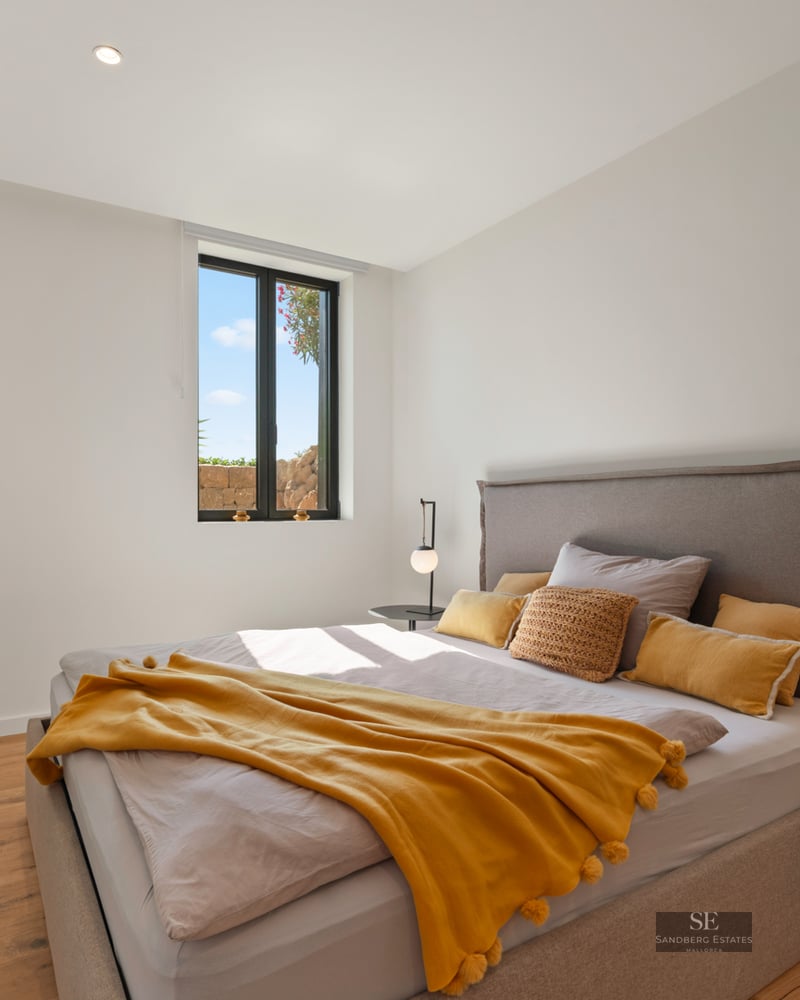 Modern bedroom with a grey bed, mustard yellow throw, wooden floors, and a window showing a bright sky.