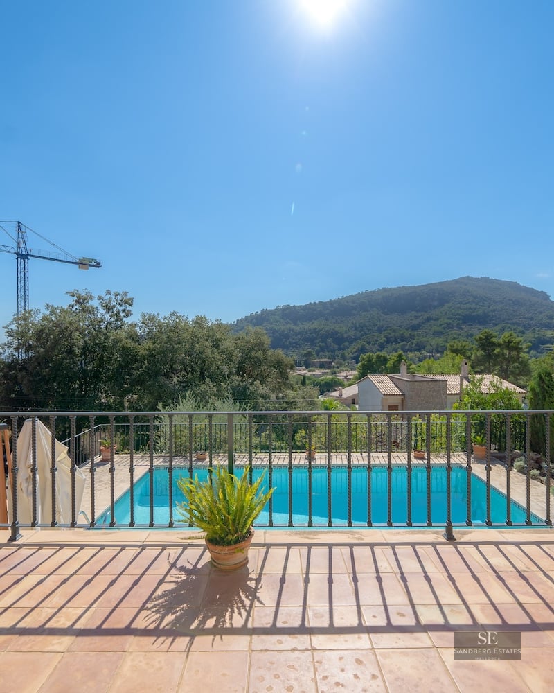 View from a terrace with iron railing overlooking a blue swimming pool and green mountains under a bright sun.