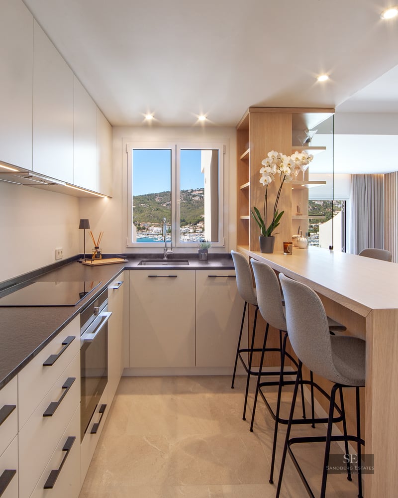 Modern kitchen with white cabinets, dark countertops, a wooden breakfast bar with three stools, and a window view of a harbor.