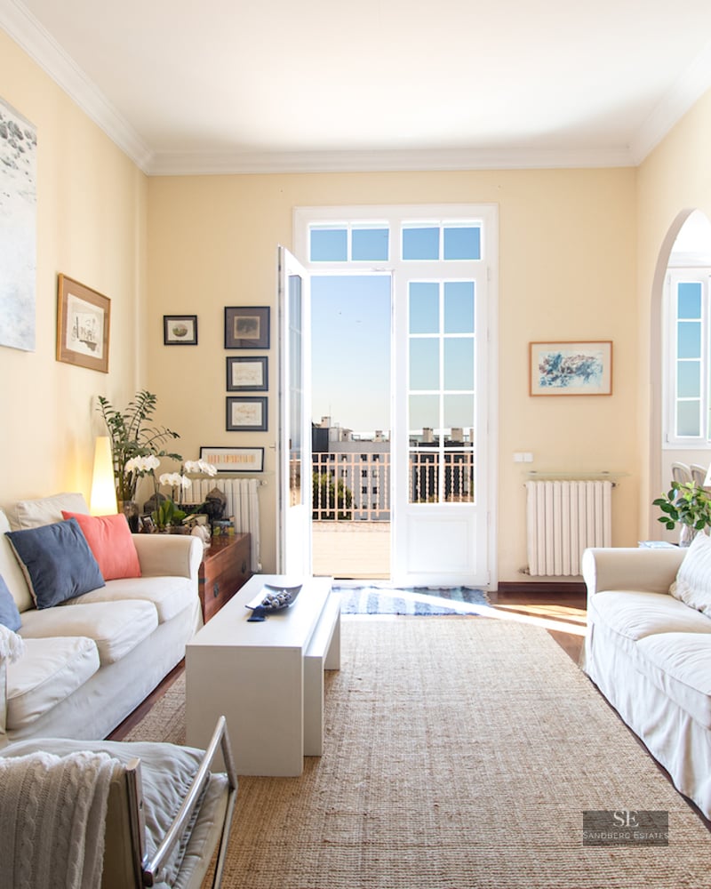 Sun-drenched living room with white sofas, a jute rug, and French doors leading to a balcony with city views.