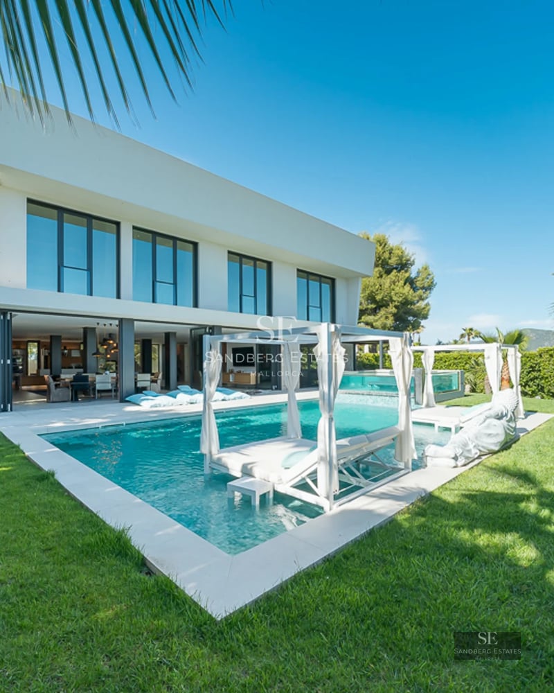 View of turquoise pool surrounded by lush greenery and modern architecture. White lounge chairs and umbrellas invite relaxation.