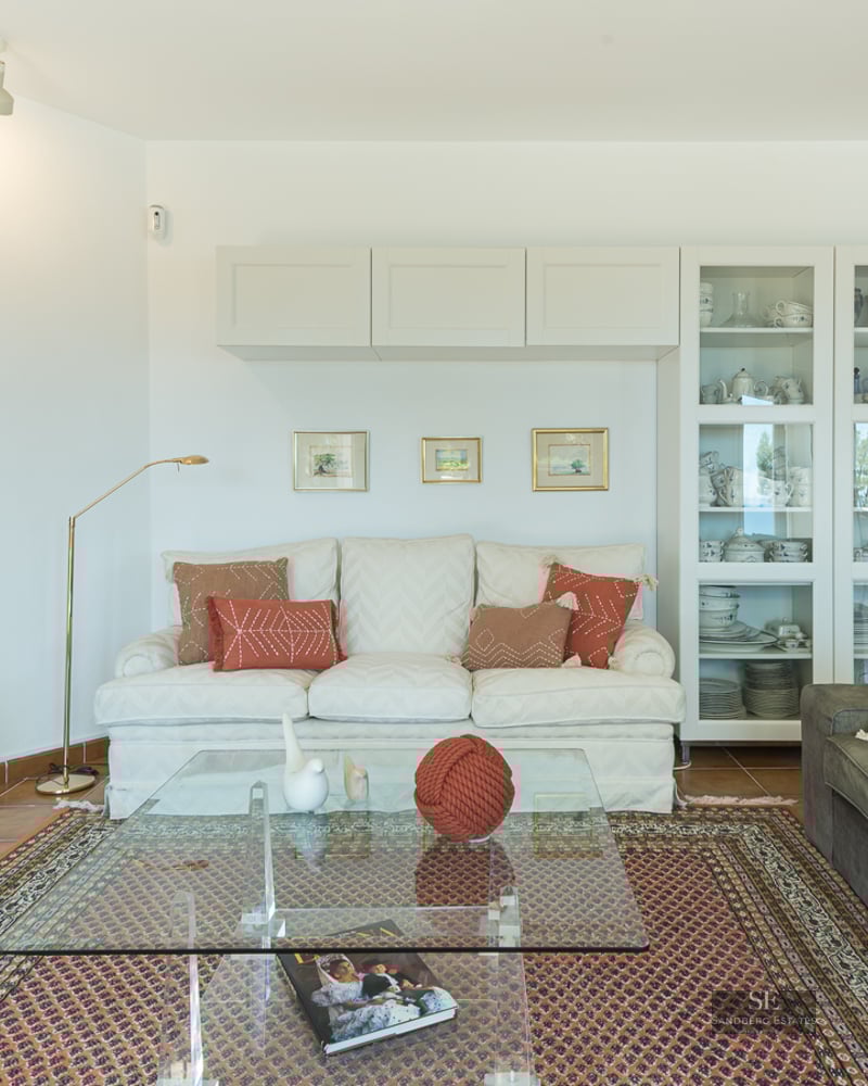 Bright living room with sofas, glass coffee table on an ornate rug, large TV, and terracotta tile floors.