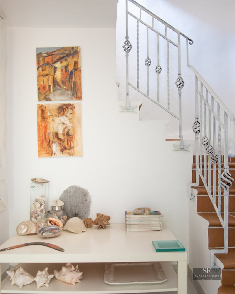 A white hallway featuring terracotta tiled stairs, a wrought iron railing, and a console table with coastal decor.