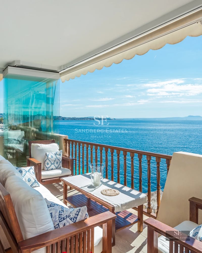 A sun-drenched terrace with wooden seating and white cushions overlooking a vast blue ocean under a clear sky.