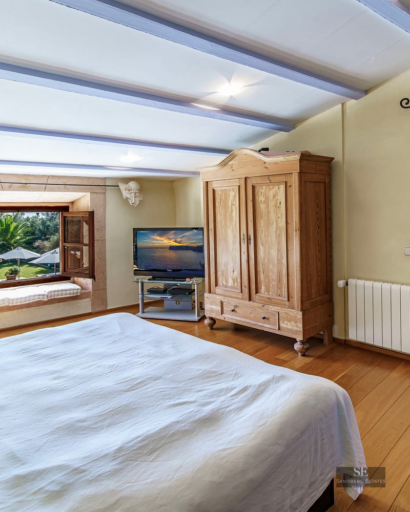 A cozy bedroom with wooden floors, a large bed, rustic armoire, and a window overlooking a lush garden.