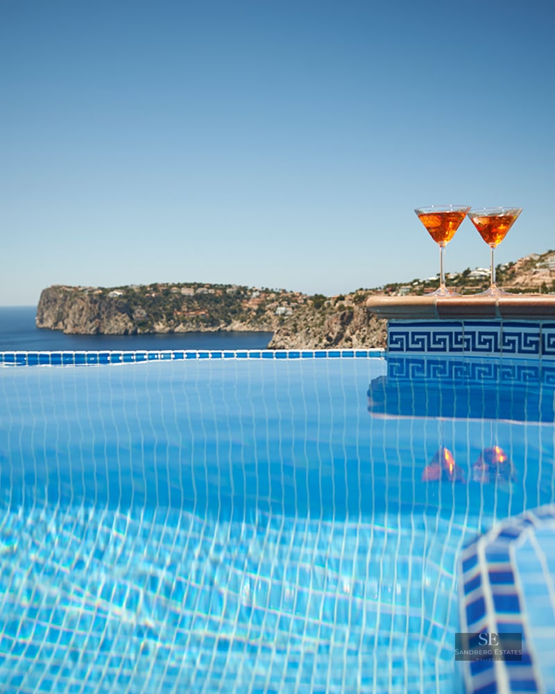 Close-up of a blue tiled infinity pool with two orange cocktails on the ledge overlooking the sea and cliffs.