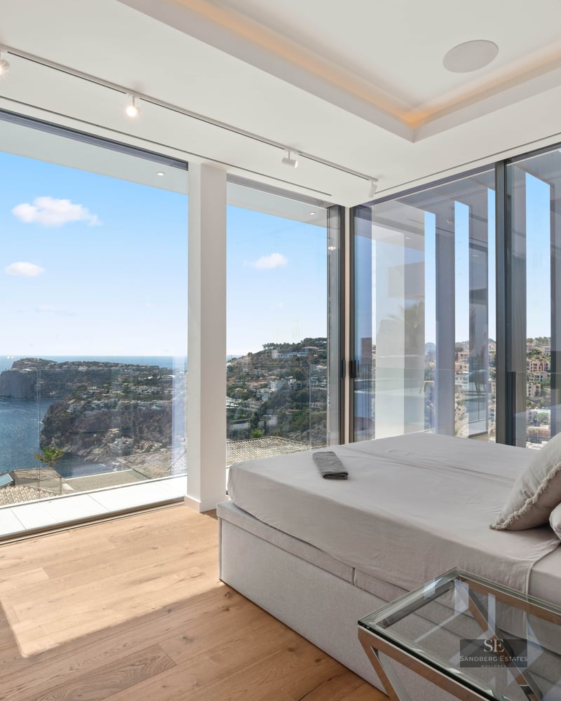 Modern bedroom with floor-to-ceiling windows showing a panoramic view of the sea and coastline.