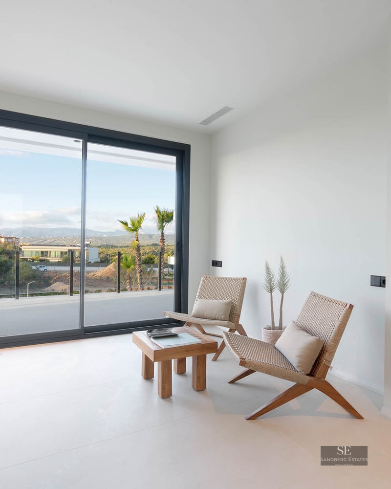 Minimalist white room featuring two design chairs, a wooden table, and large sliding glass doors overlooking a landscape.