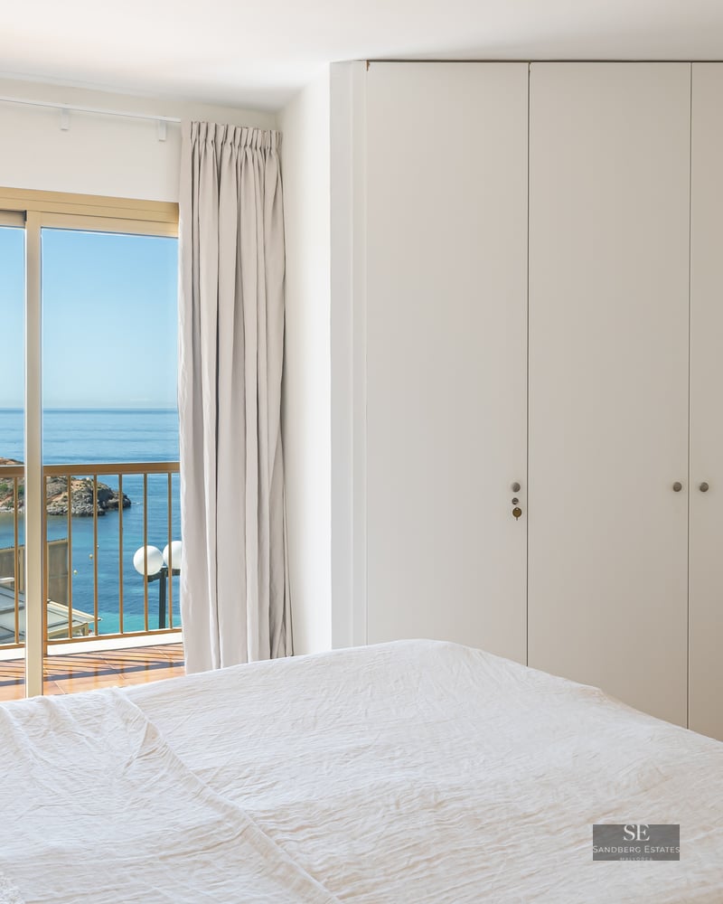 Bright bedroom with white bedding, wooden crib, built-in wardrobe, and sliding glass door opening to a sea view.