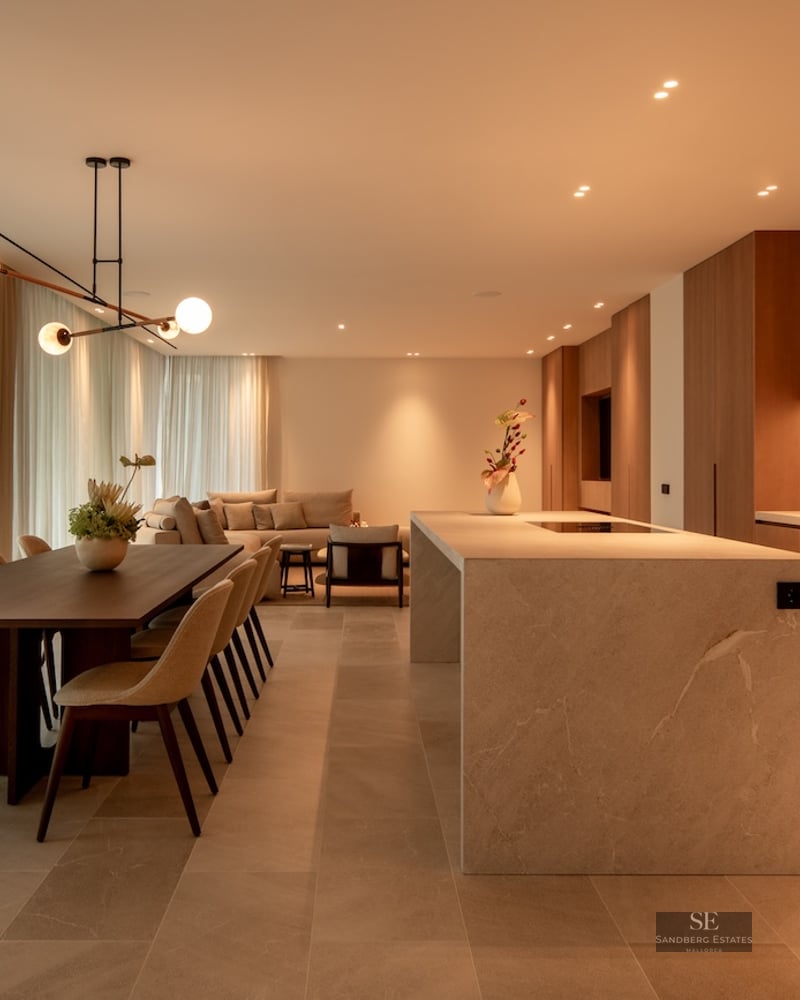 Warmly lit open-plan room with a stone kitchen island, dining table, and sofa behind soft curtains.