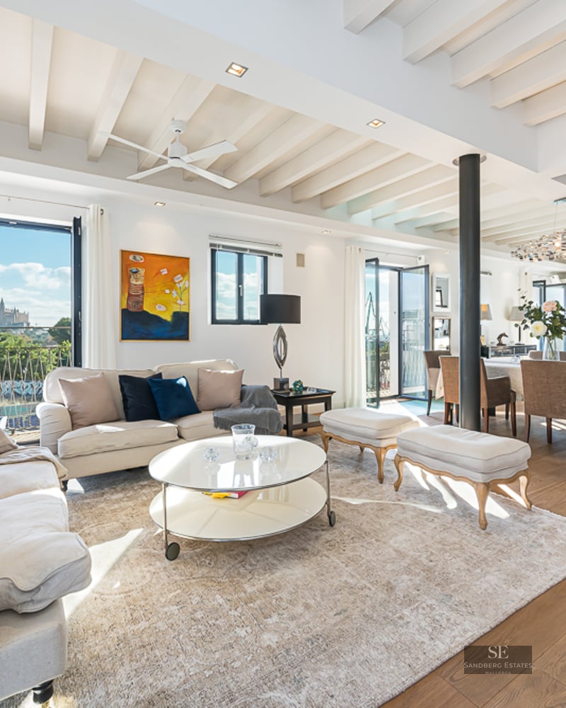 Spacious white living room with beamed ceilings, wooden floors, and a view of a cathedral through large windows.