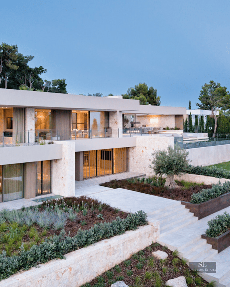 Modern multi-level villa with stone walls, large windows, and tiered Mediterranean gardens overlooking the sea at dusk.