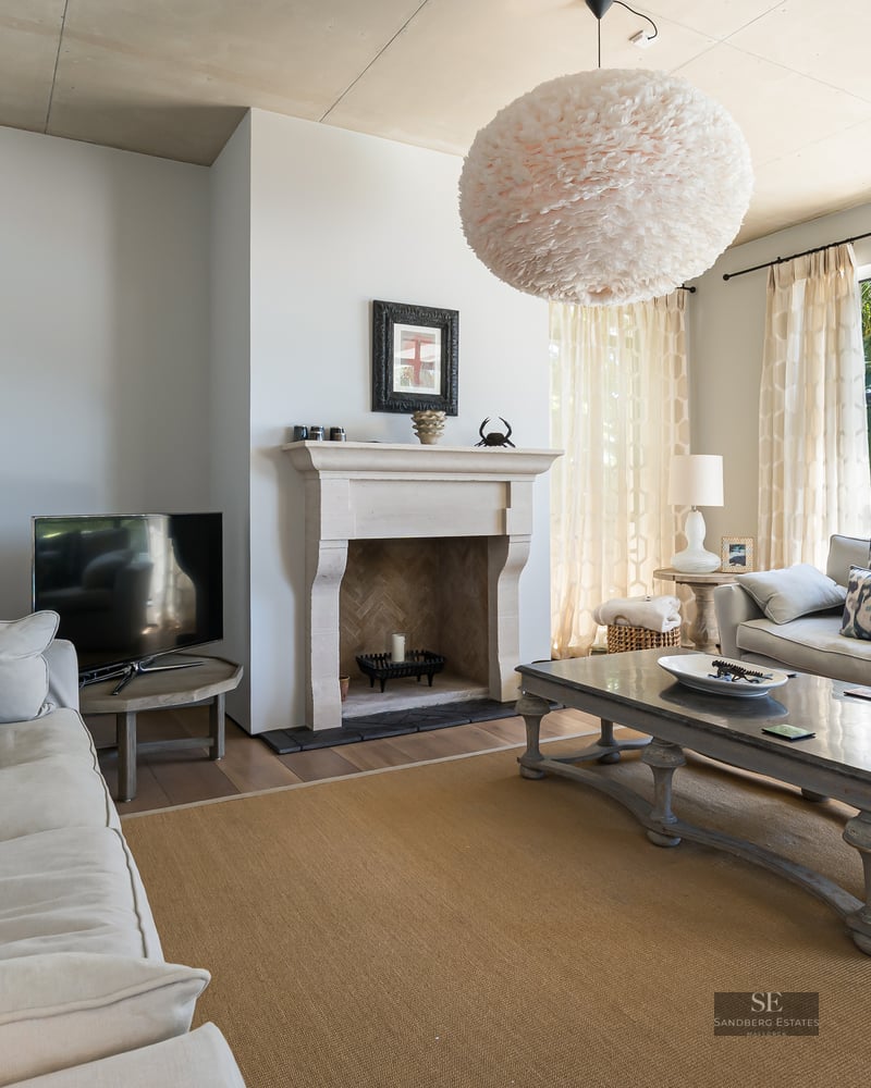 Spacious living room with beige sofas, stone fireplace, and large windows viewing garden palms.