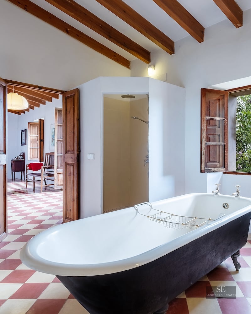 Spacious bathroom featuring a black freestanding clawfoot tub, exposed wooden beams, checkered tile floor, and garden view.
