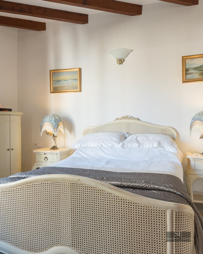 Cozy bedroom featuring a rattan bed, wooden ceiling beams, bedside lamps, and dark wood window shutters.
