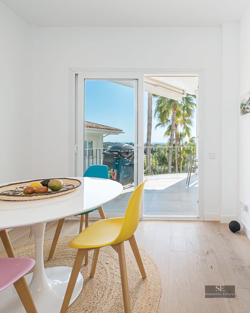 Salle à manger moderne avec table blanche et chaises colorées près de baies vitrées donnant sur une terrasse.