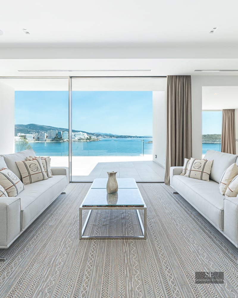 Bright living room with two grey sofas, a glass coffee table, and floor-to-ceiling windows overlooking the ocean.