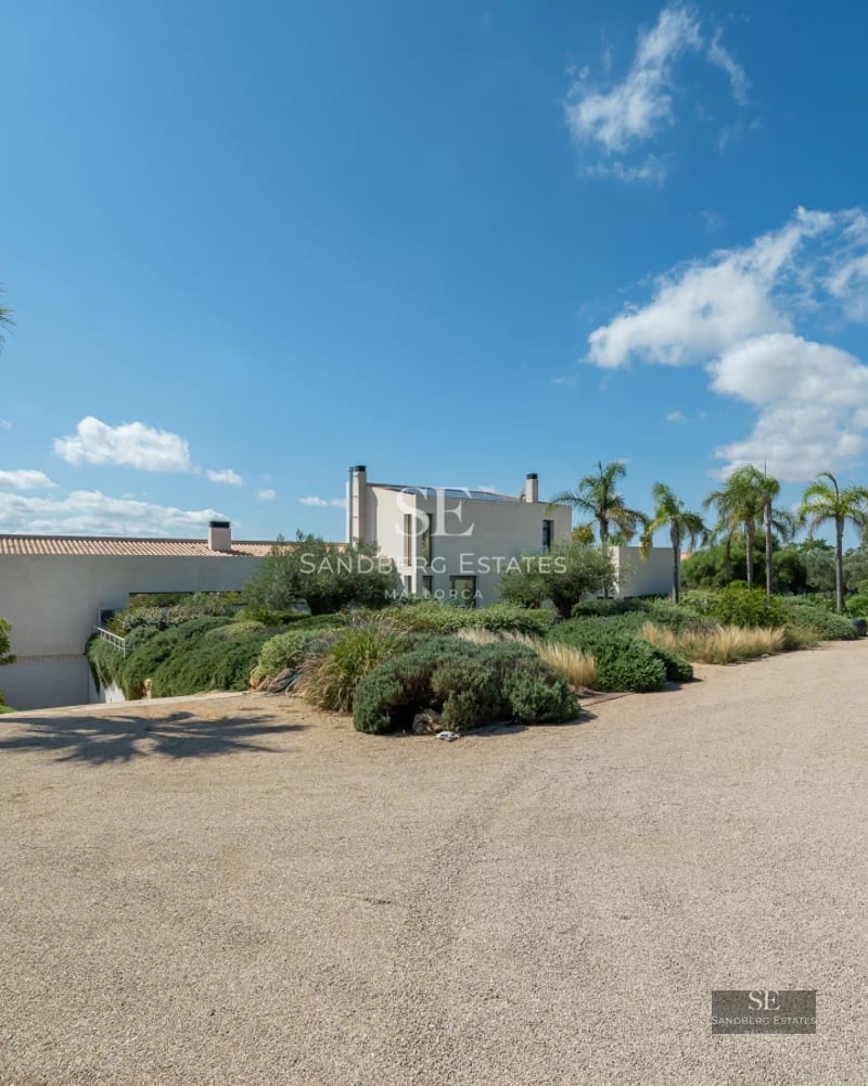View of infinity pool surrounded by a modern villa with large windows and minimalist design. Well-manicured garden and blue sky.