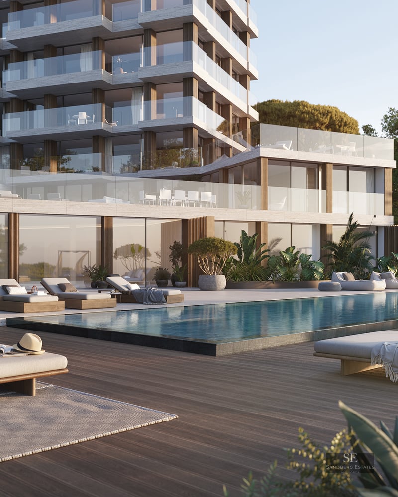 Modern apartment building with a large swimming pool, wooden deck, luxury lounge chairs, and lush green gardens.