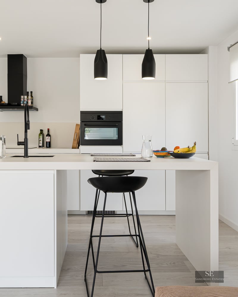 Bright white kitchen featuring a large central island with black bar stools and black hanging pendant lights.