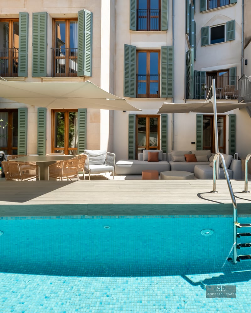 A turquoise swimming pool with a wooden deck, lounge furniture under shade sails, and a building with green shutters.