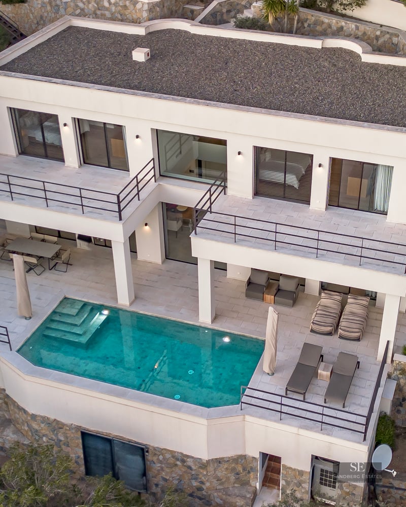 Aerial view of a modern white villa featuring a turquoise pool, multi-level terraces, and cypress trees.