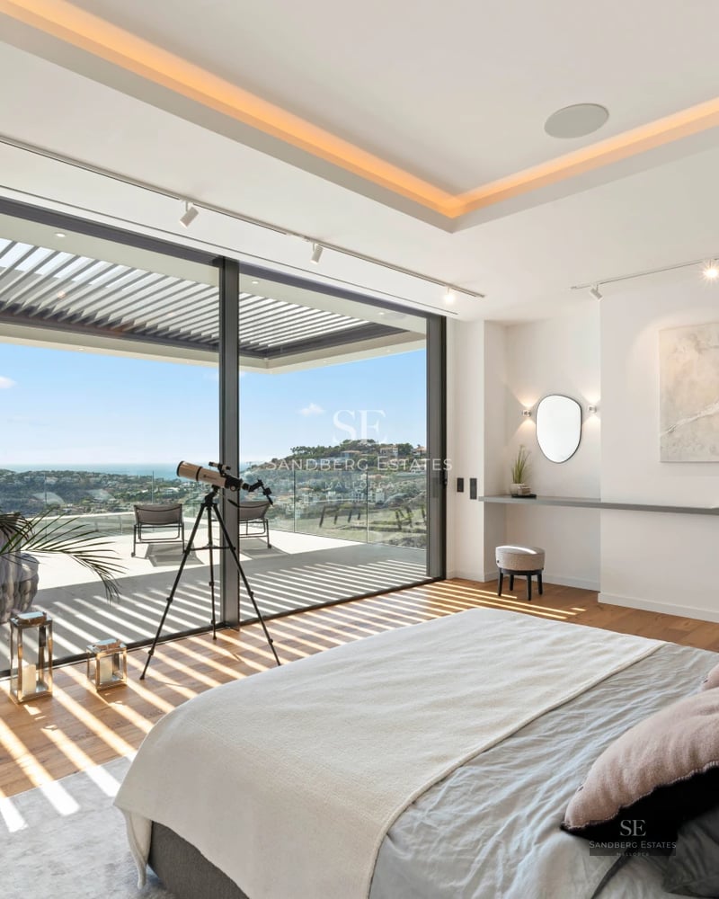 View of a turquoise pool surrounded by greenery and a modern house with large windows reflecting the blue sky.