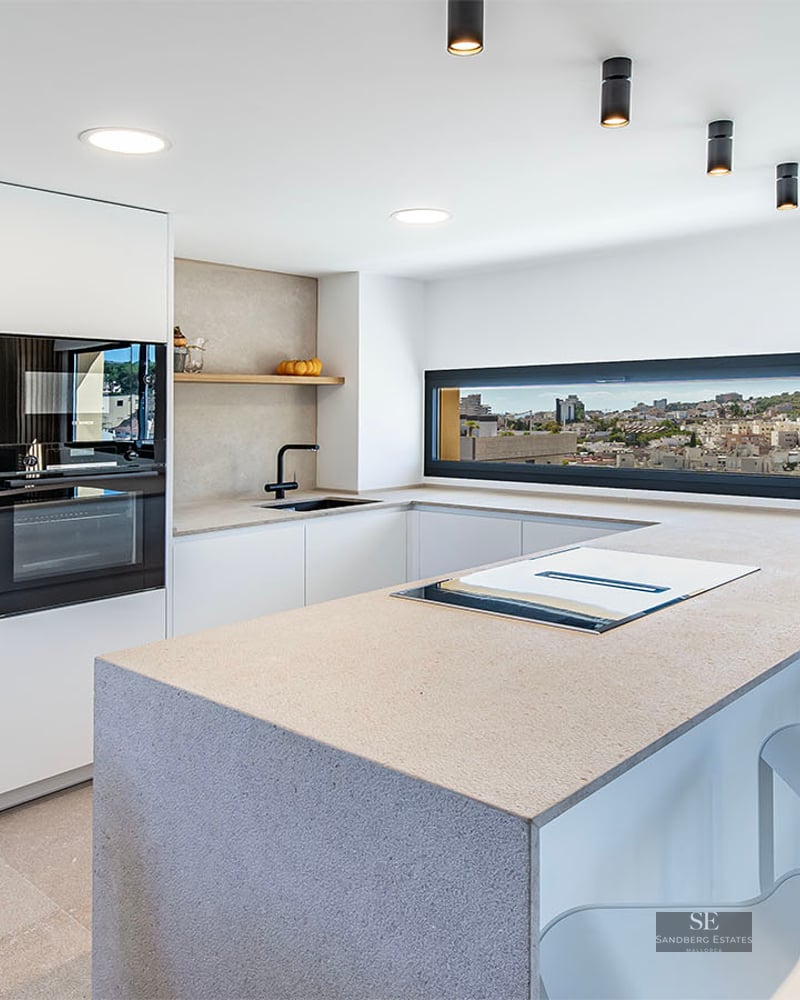 Minimalist white kitchen with large stone island, integrated appliances, and a panoramic window overlooking a city.