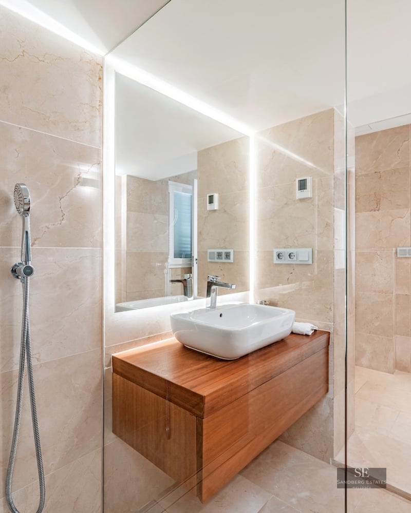 Modern bathroom featuring beige marble walls, a glass walk-in shower, and a floating wood vanity with a white vessel sink.