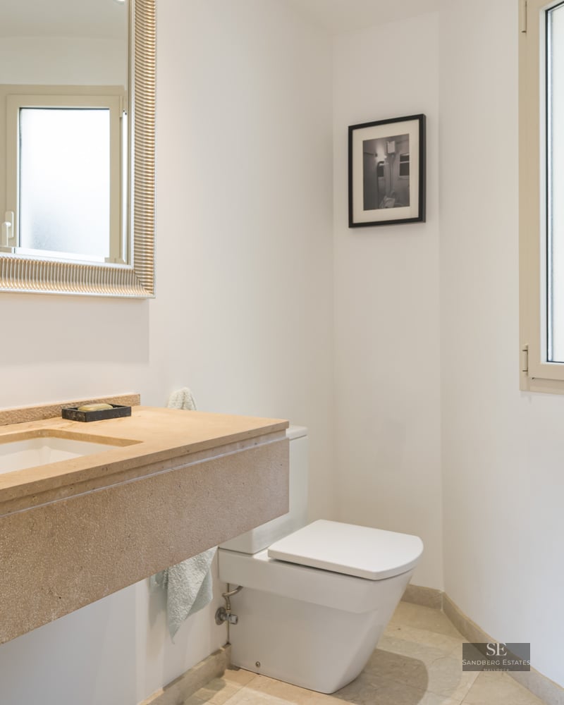 Bright bathroom featuring a tan stone vanity, large silver-framed mirror, white toilet, and frosted window.