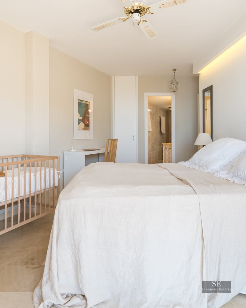 Bright bedroom featuring a large bed, wooden baby crib, workspace, and polished marble flooring.