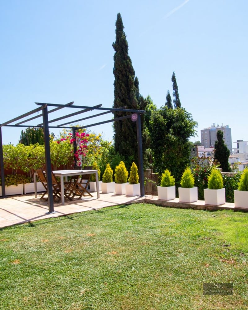 Sunny terrace with a dining table under a metal pergola, a lawn area, and white planters overlooking a city view.