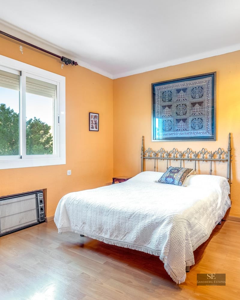 Bedroom with orange walls, white bed, ornate metal headboard, large wooden wardrobe, and window view of trees.