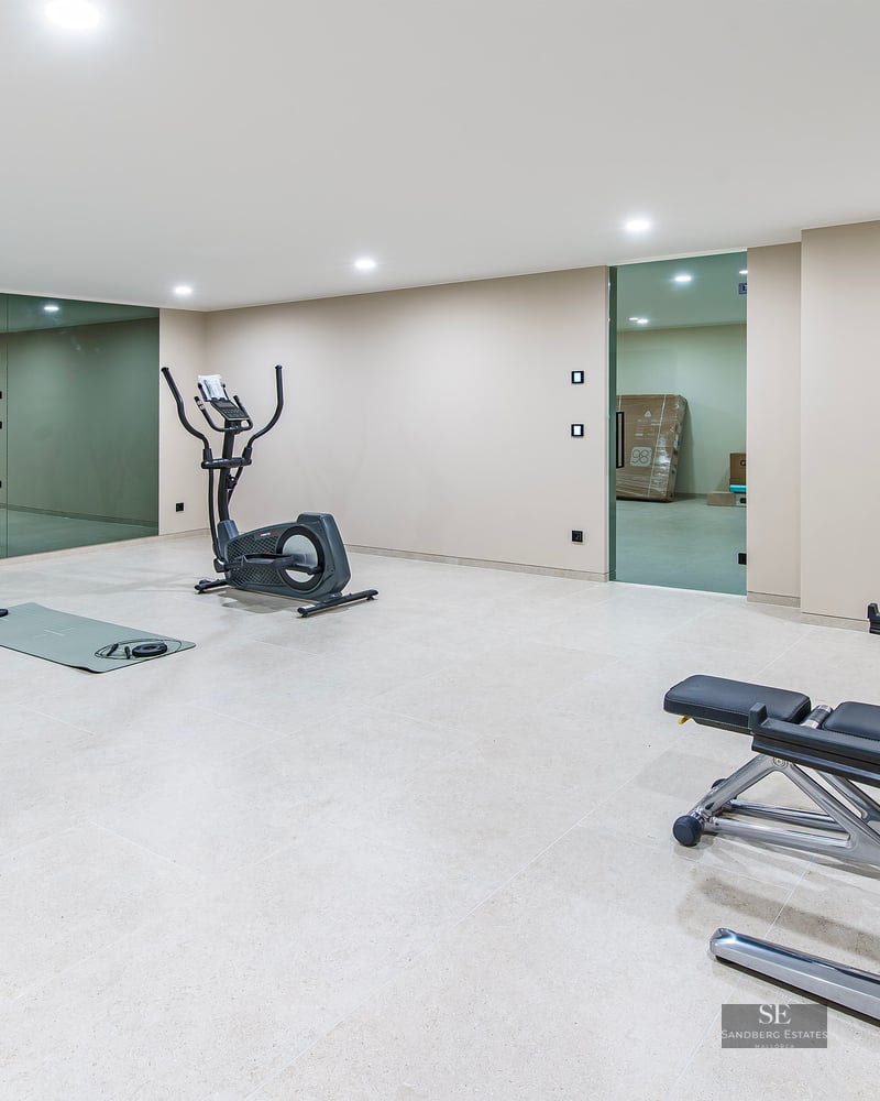 Spacious gym with elliptical trainer, weight bench, mats, and large wall mirrors under bright ceiling lights.