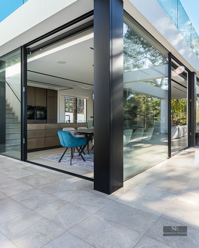 Luxury terrace with large floor-to-ceiling glass windows, light stone flooring, and a view into a modern interior kitchen.