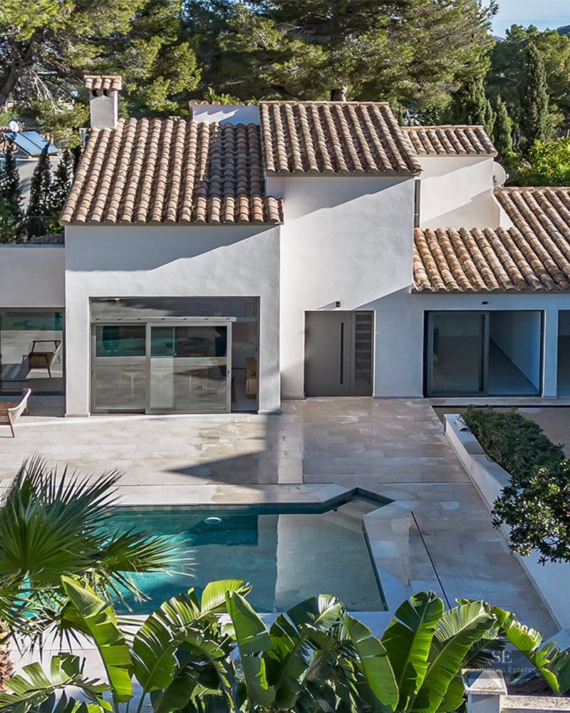 Aerial view of a white villa with a terracotta roof, a turquoise swimming pool, and stone terrace surrounded by pine trees.