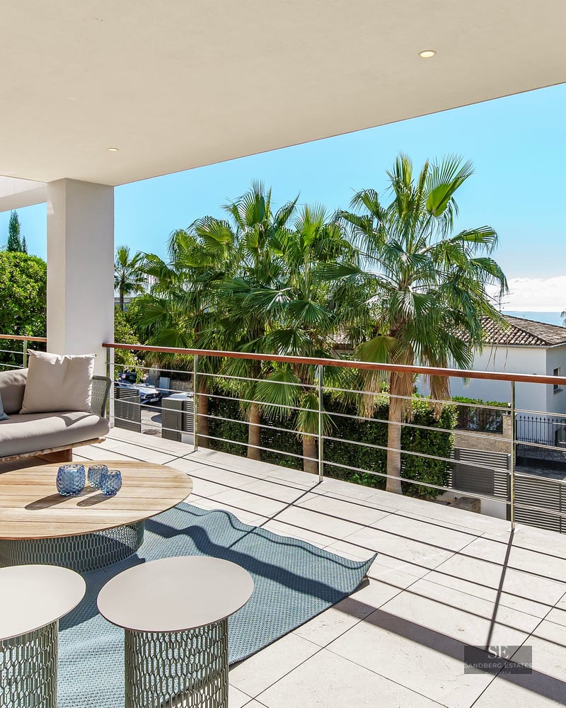 Terrasse moderne avec canapés gris, table en bois et touches turquoise offrant une vue sur les palmiers et la mer.
