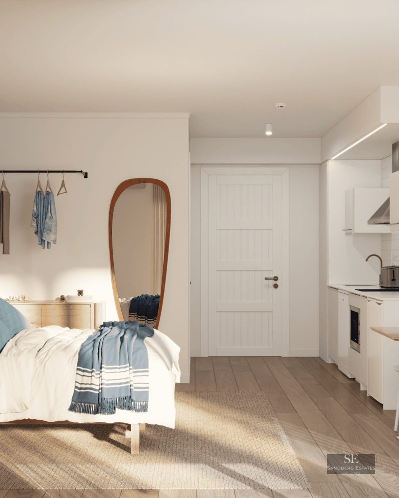 Modern studio with bed, white kitchenette, small dining table, and natural light on light wood floors.