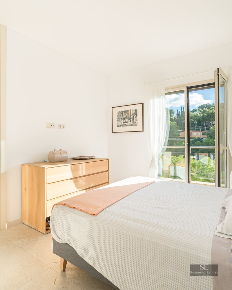 Bright bedroom featuring a bed, light wood dresser, and open balcony overlooking lush greenery.
