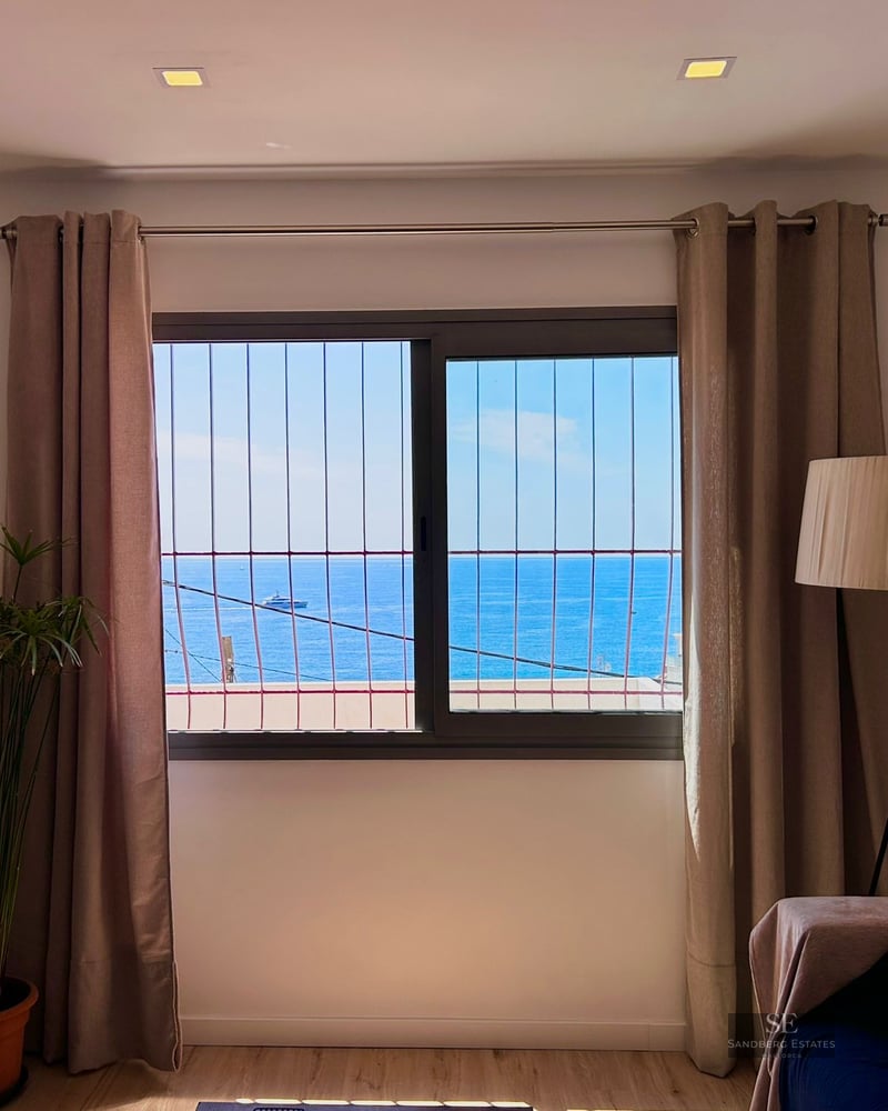 A window with a metal grate framing a blue sea view, flanked by beige curtains, a floor lamp, and a houseplant.
