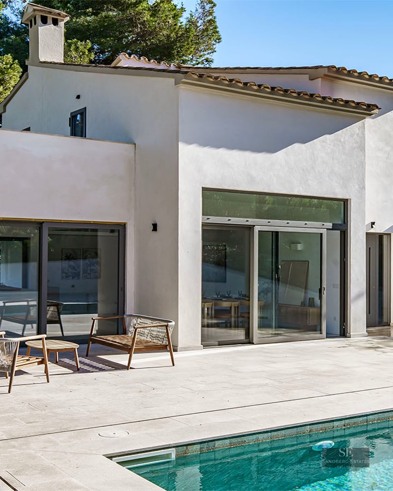A large turquoise swimming pool in front of a modern white villa with stone terrace and lounge furniture.