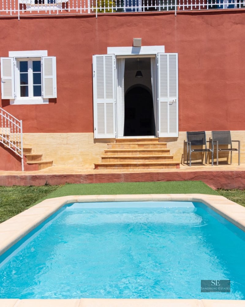 Rectangular turquoise pool in front of a red Mediterranean villa with white shutters and stone terrace.