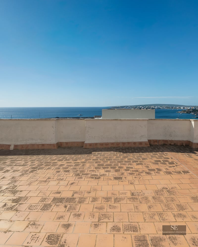 Large rooftop terrace with terracotta tiles overlooking a clear blue sea and coastline under a bright sky.